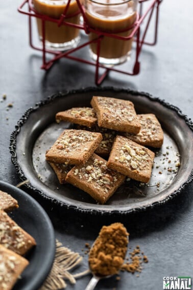 almond jaggery cookies placed in a silver plate with glasses of chai in the background
