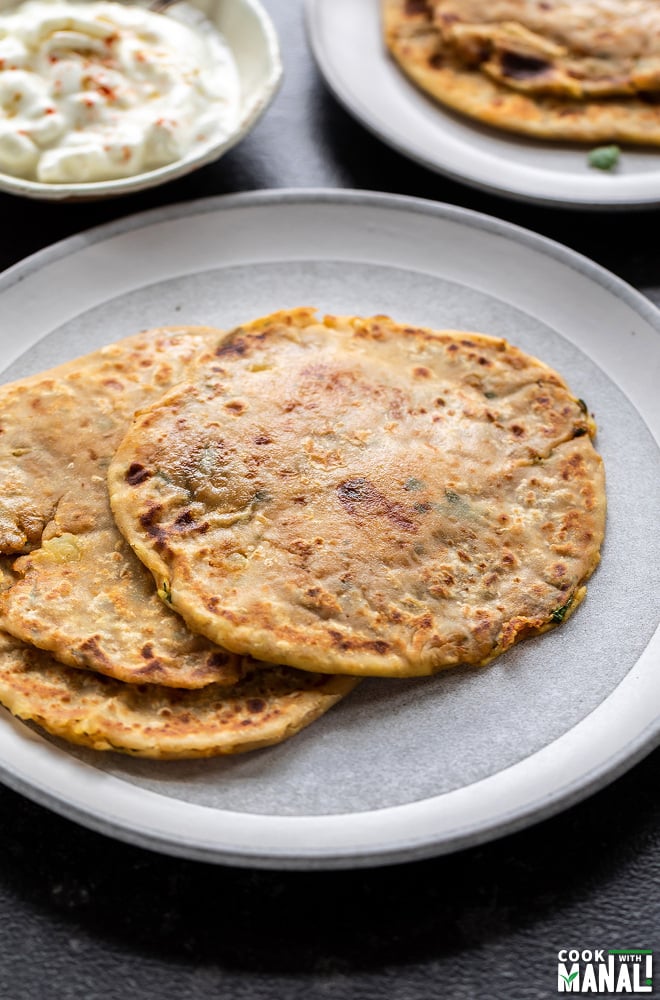 3 aloo parathas on a white plate with a bowl of yogurt in the back