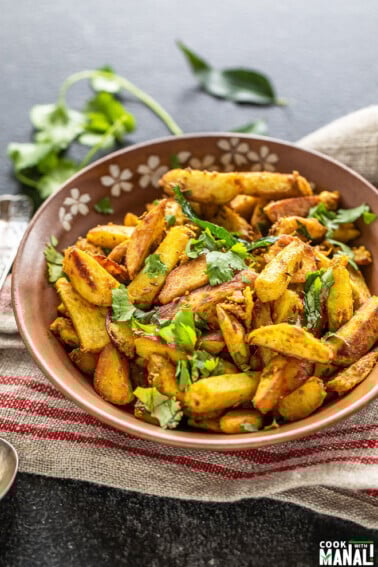 spiced arbi fry served in a brown bowl, garnished with cilantro and a twig of cilantro and curry leaves placed in the background
