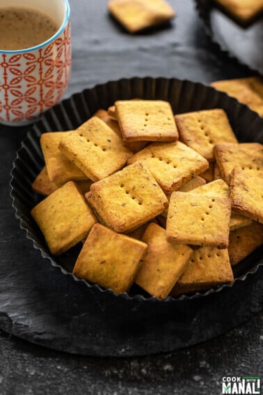 baked besan papdi in a black plate with a cup of coffee in the background