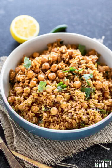 brown rice chana pulao served in a blue bowl, garnished with cilantro and a cut lemon and green chili in the background