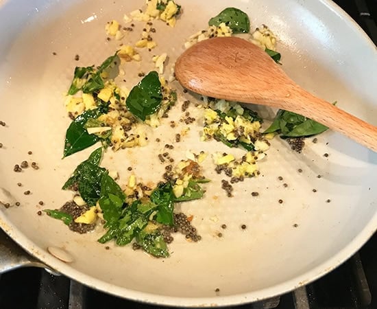curry leaves with mustard seeds being stirred with a wooden spatula in a pan