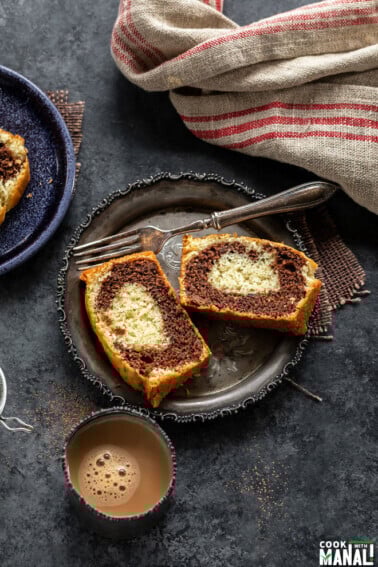 two slices of cake arranged in a plate with a fork and a cup of chai placed on the side