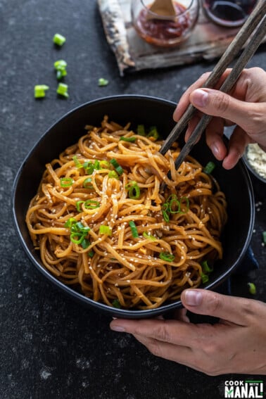 pair of hands holding a bowl of noodles with one hand holding a pair of chopsticks