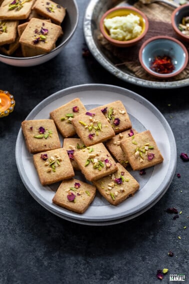 cookies topped with rose petals and pistachios placed on a plate with small bowls containing saffron placed in the background