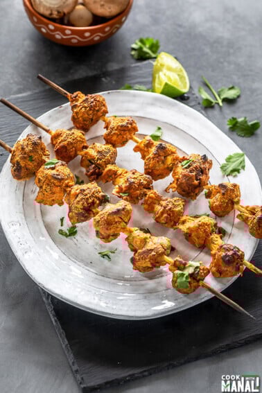three wooden skewers of mushroom tikka arranged on a white plate and garnished with cilantro
