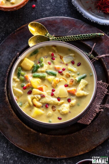 yellow color vegetable curry in a round bowl garnished with pomegranate arils and two golden color spoons placed in the background