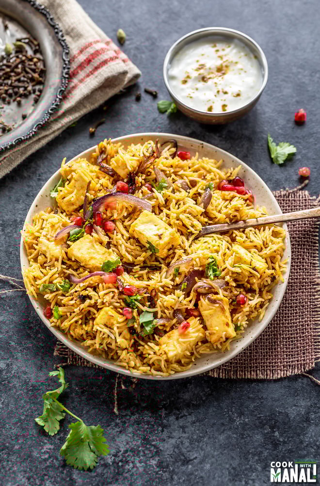 a plate full of rice and paneer, garnished with cilantro and pomegranate arils with a bowl of yogurt place on one side and a plate of spices placed on the other side