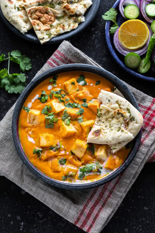 bowl of paneer butter masala served with a piece of naan and garnished with cilantro and cream