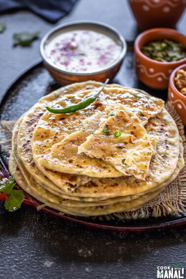 stack of paneer paratha with green chili on the side and bowls of yogurt, pickle in the background