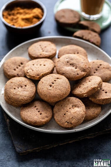 plate full of ragi cookies with bowl of jaggery and a glass of chai in the background
