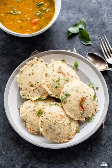 5 pieces of rava idli arranged in a plate with a bowl of sambar in the background