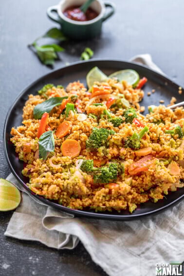 black plate with quinoa, veggies, basil, lime wedge and more basil and a bowl of hot sauce in the background