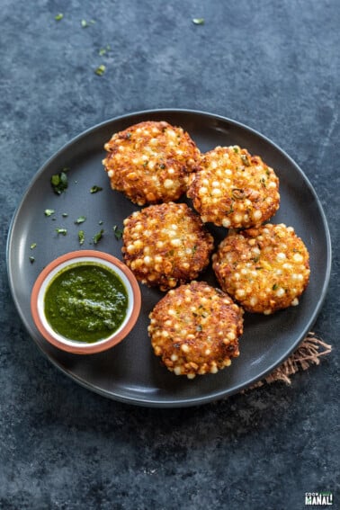 5 pieces of sabudana vada arranged in a plate with a small bowl of cilantro chutney on the side